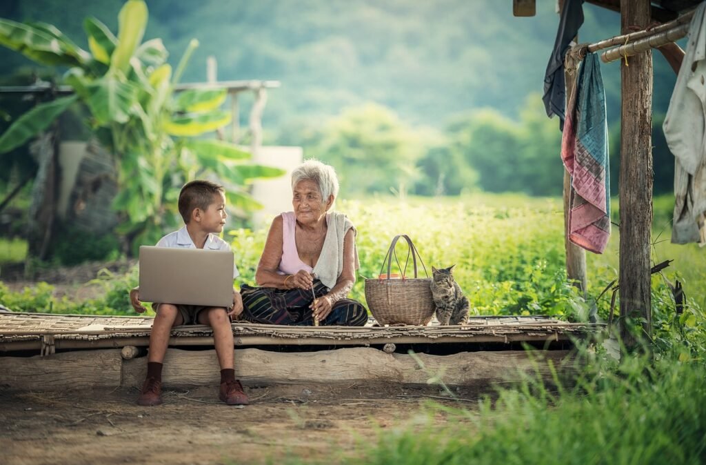 An elderly woman and a young boy sit together on a wooden bench, engaged with a laptop in a serene outdoor setting.